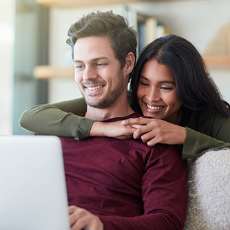 Foto de mulher jovem abraçada ao homem, ammbos sorrindo e olhando para a tela do laptop